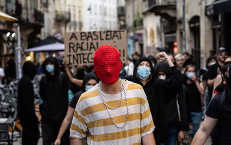 Photo of the anti-racism protests in reaction to the murder of the Black teenager Nahel in France. While most people are dressed in normal riot gear, with black hoodies and COVID masks, the photo focus on a protester who is wearing a red net over their had that covers their entire face, eyes and nose and all.  Underneath they wear a striped yellow-white T-shirt and large, hip-hop style chains.