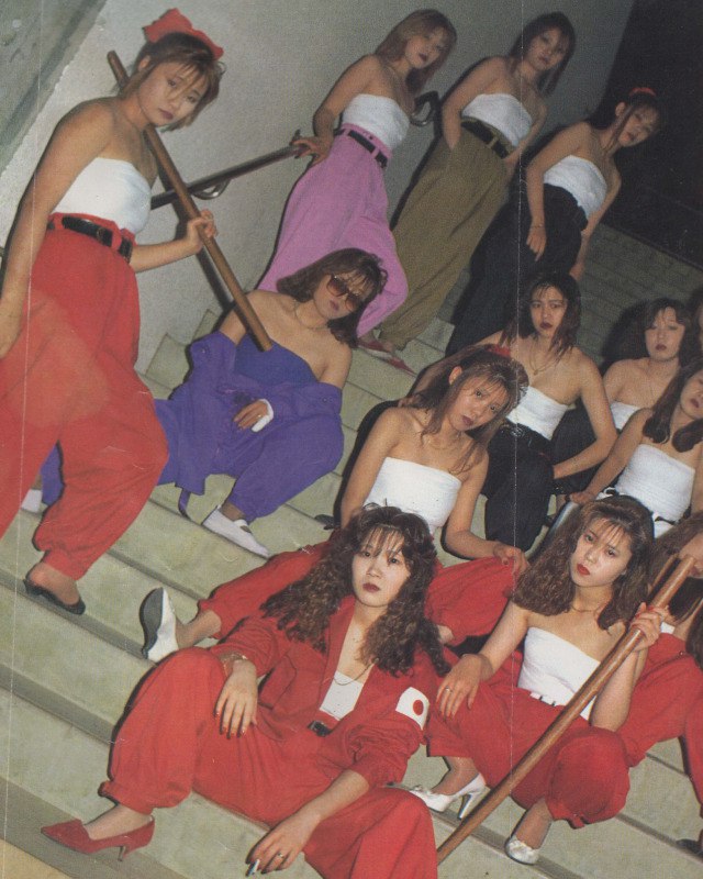 Young delinquent biker women in Japan posing in a staircase.  Most of them have colourful wide pants, cotton bandages binding their breasts, long hair bleached brown and often wavy with perms, and ooze confidence and style.  Many of them wield wooden swords menacingly for the camera.